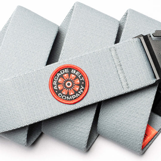 A light gray Arcade Belt Co. from Arcade Belts, made of stretch webbing with a black buckle and a red-and-black circular embroidered patch. Perfect for travel belt enthusiasts.