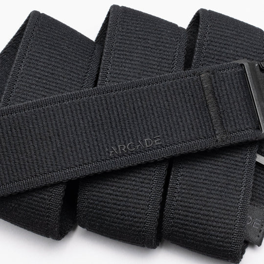 A close-up of the Atlas belt by Arcade Belts shows its black, textured design with ARCADE embossed on it. The belt is neatly folded, displaying a sleek plastic buckle partially visible on the right side.
