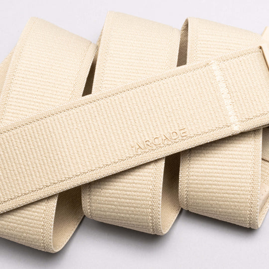 A close-up of the Atlas belt by Arcade Belts, featuring beige elastic material embossed with ARCADE, arranged in overlapping loops on a light background.
