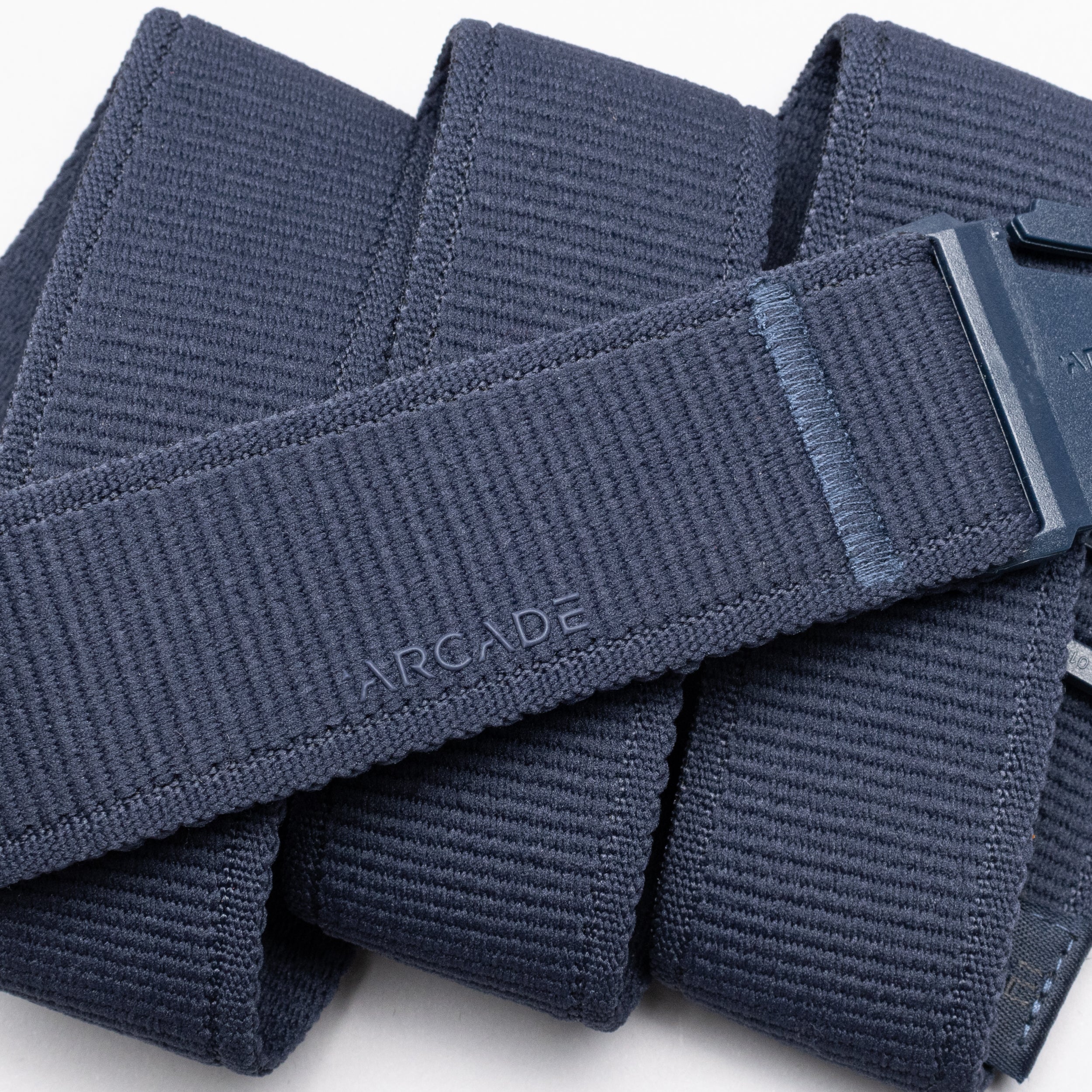 A close-up image features the Atlas belt by Arcade Belts, a navy blue elastic stretch belt with a plastic buckle and subtle ARCADE branding. Neatly folded, it’s perfect for travelers seeking comfort and a low-profile design.