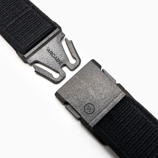 Close-up of the Atlas belt by Arcade Belts, a black elastic stretch belt with a metal quick-release buckle—partially unclasped, ARCADE engraved on one side. White background.