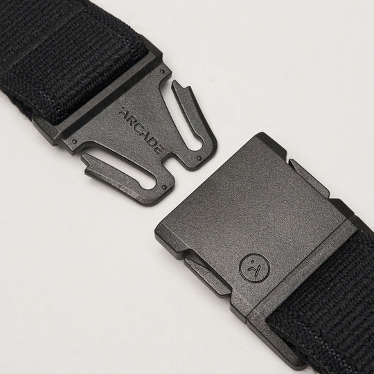 A close-up of the Atlas elastic stretch belt by Arcade Belts in black, featuring a plastic quick-release buckle. The unfastened low-profile buckle shows the Arcade Belts brand name on one side and a small circular face logo on the other.