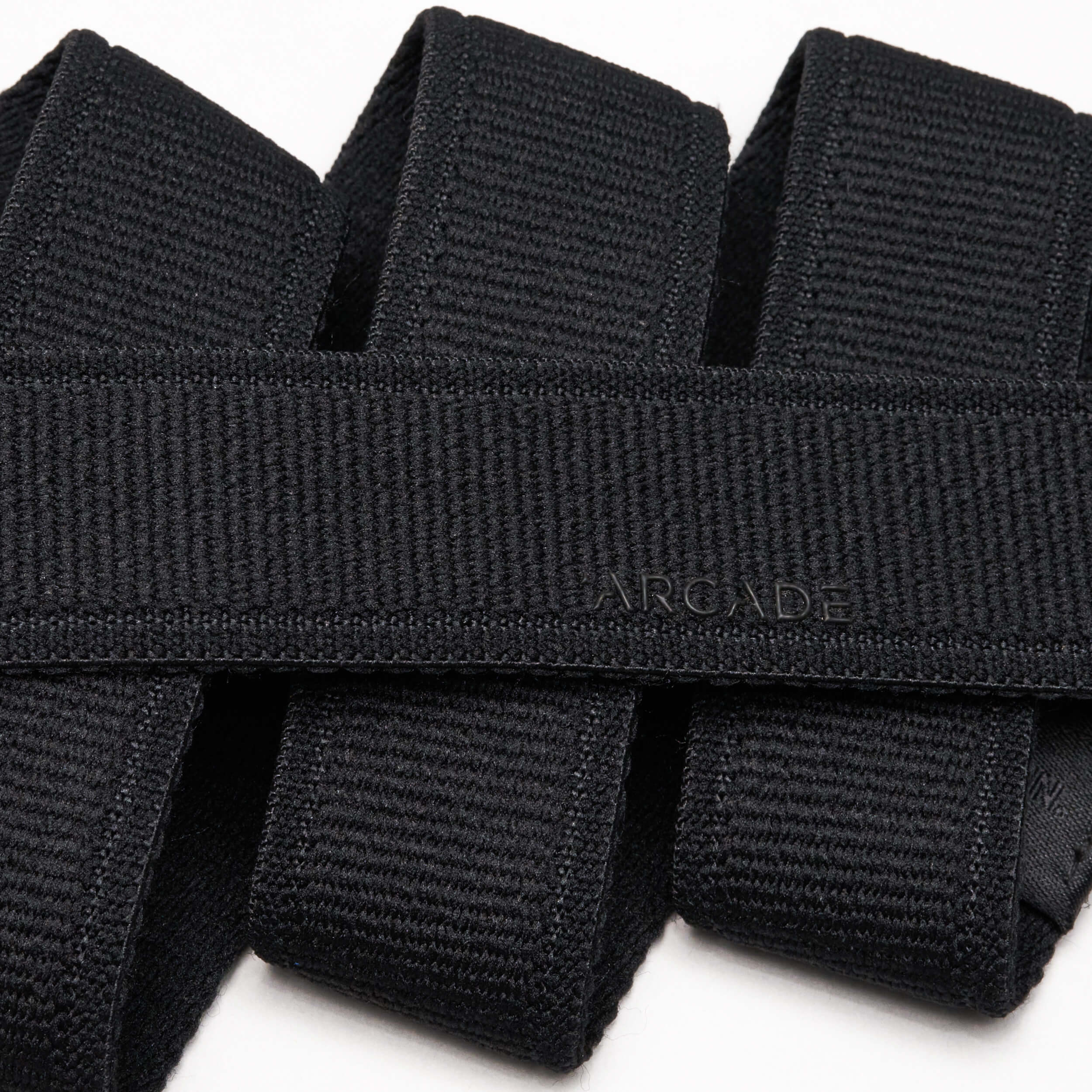 A close-up photo of overlapping black Atlas belts from Arcade Belts, with the word ARCADE subtly embossed on one of the elastic stretch belts near the center.