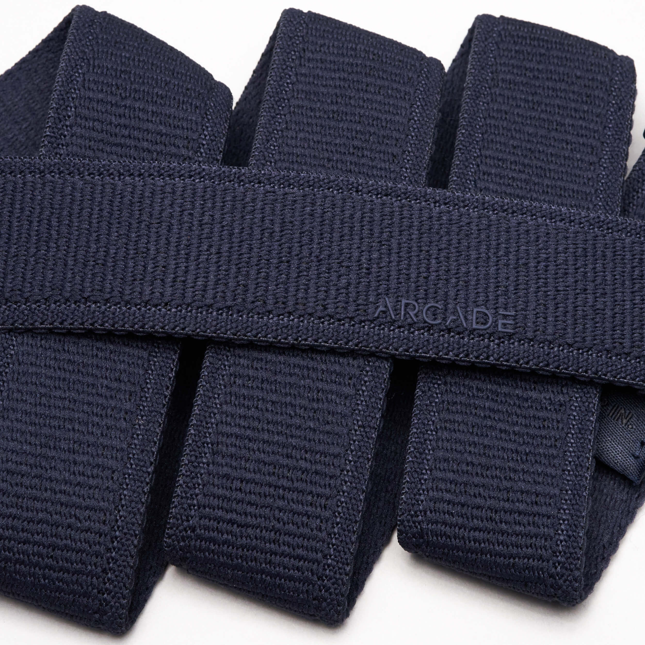 Close-up of several folded navy blue Atlas stretch webbing belts by Arcade Belts stacked neatly, with one low profile belt subtly embossed with ARCADE on its surface.