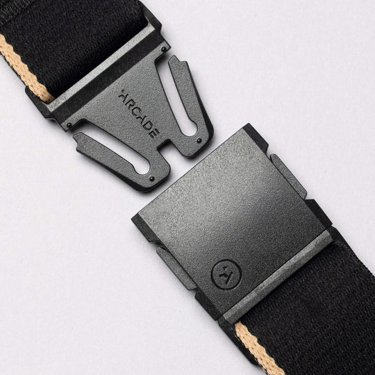 Close-up of the Arcade Belts Blackwood: a black plastic A2 buckle with ARCADE embossed, partially unclasped, attached to a black strap with beige stripe—evoking Blackwood trees—set against a light gray background.