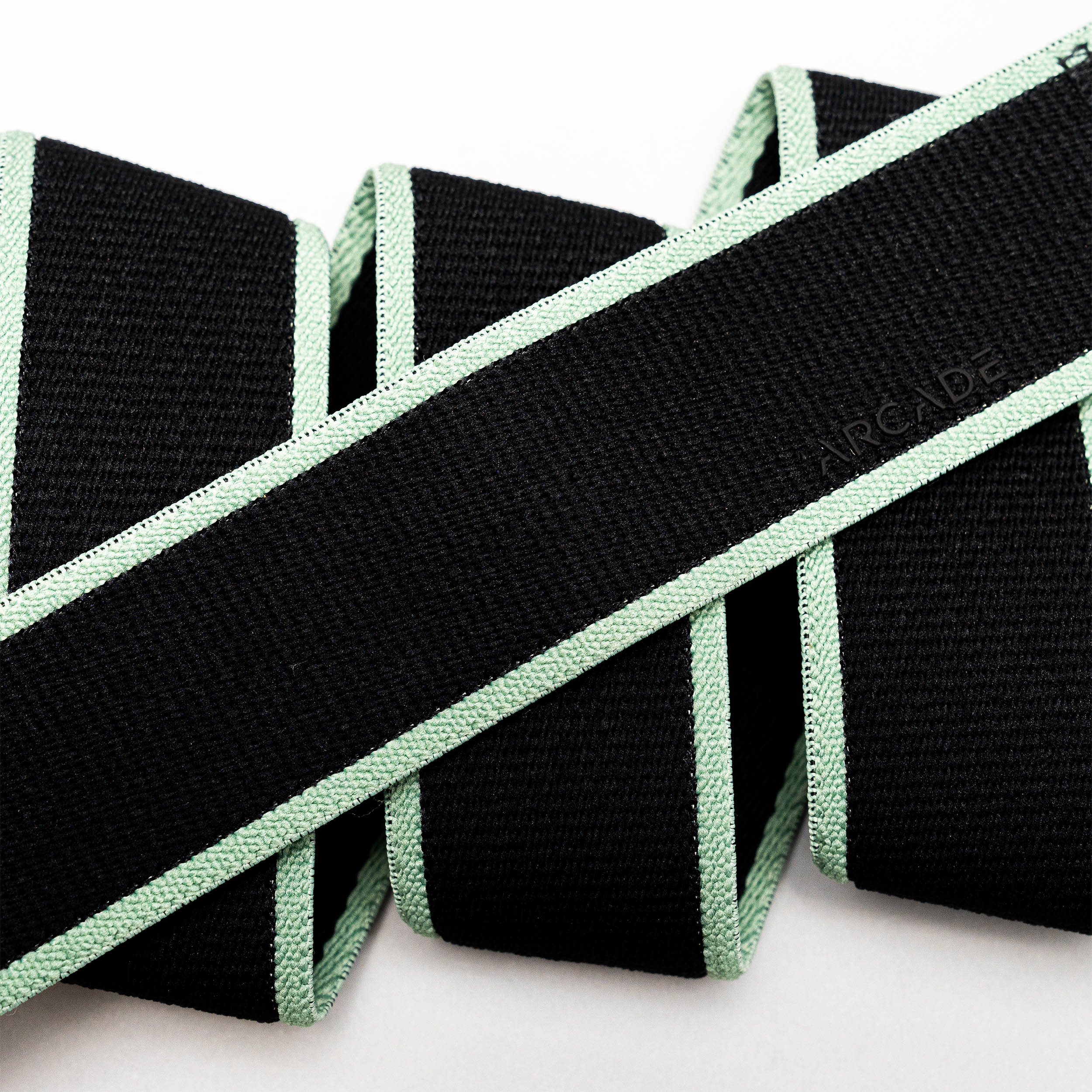 A close-up of the Arcade Belts Carto, a black stretch belt with light green edges, folded in a zigzag on a white background. The word ARCADE is subtly embossed on the webbing.