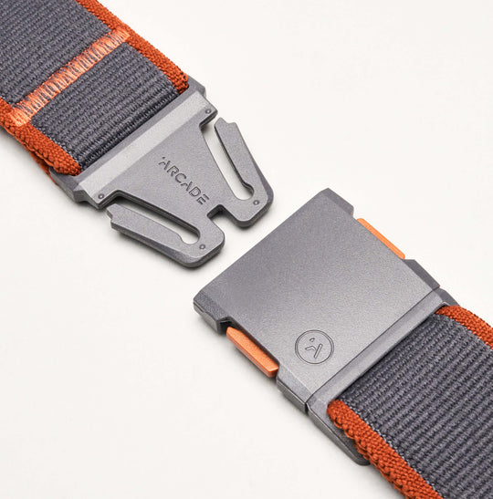 Close-up of a grey and orange Carto belt by Arcade Belts, made from nylon elastic stretch material with a metal magnetic buckle featuring the brand name and logo, partially fastened on a white background.