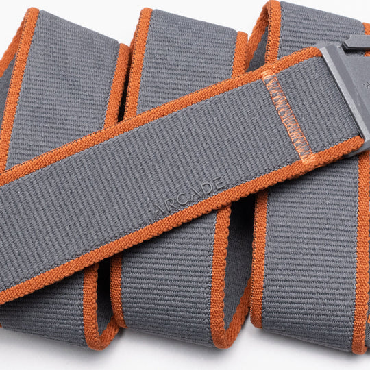 A close-up of the Carto, a gray elastic stretch belt by Arcade Belts, featuring orange edges and a rectangular plastic buckle. ARCADE is embossed on the coiled belt, highlighting its texture and stitching—perfect for travel belt lovers.
