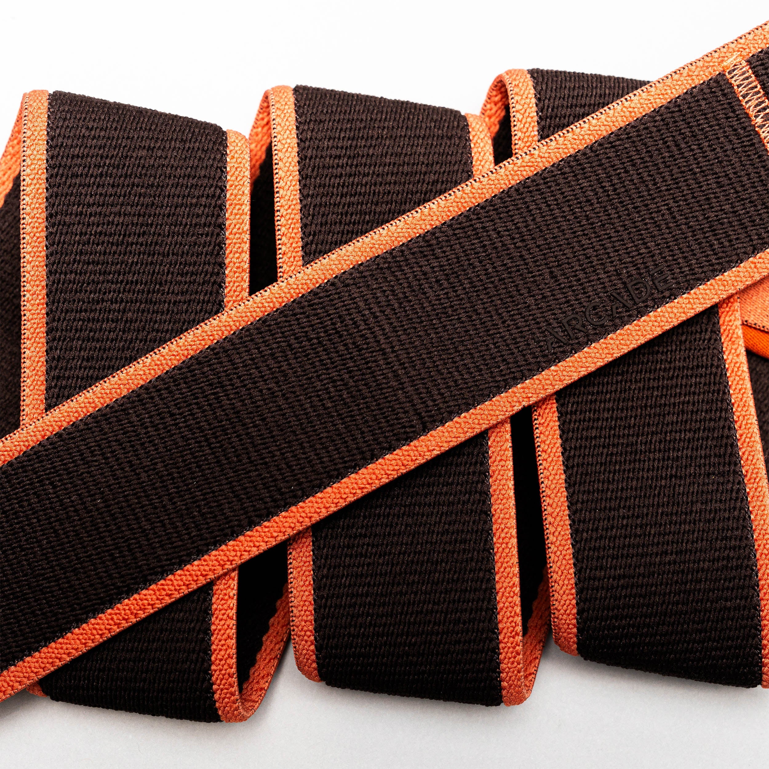 A close-up of Carto by Arcade Belts reveals brown textured straps with bright orange edges, overlapping on a white background to highlight the versatility of their stretch webbing.
