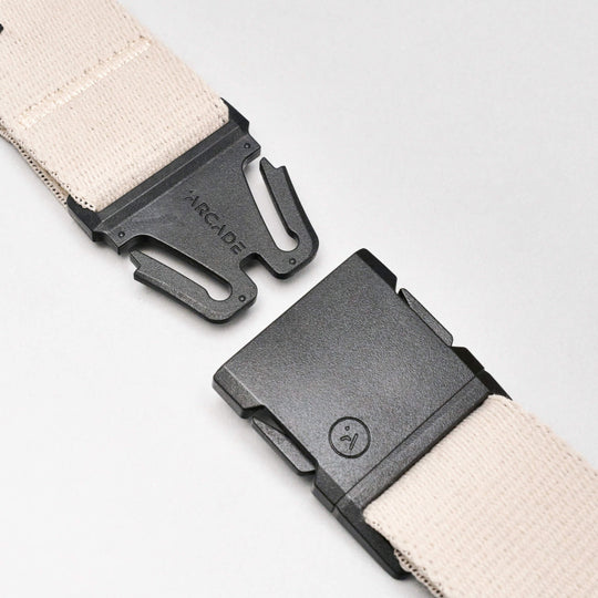 A close-up of the beige Charmer Slim travel belt by Arcade Belts, featuring a black plastic buckle partially unclasped to show the interlocking parts on a plain background. The Arcade logo appears on one side of the buckle.