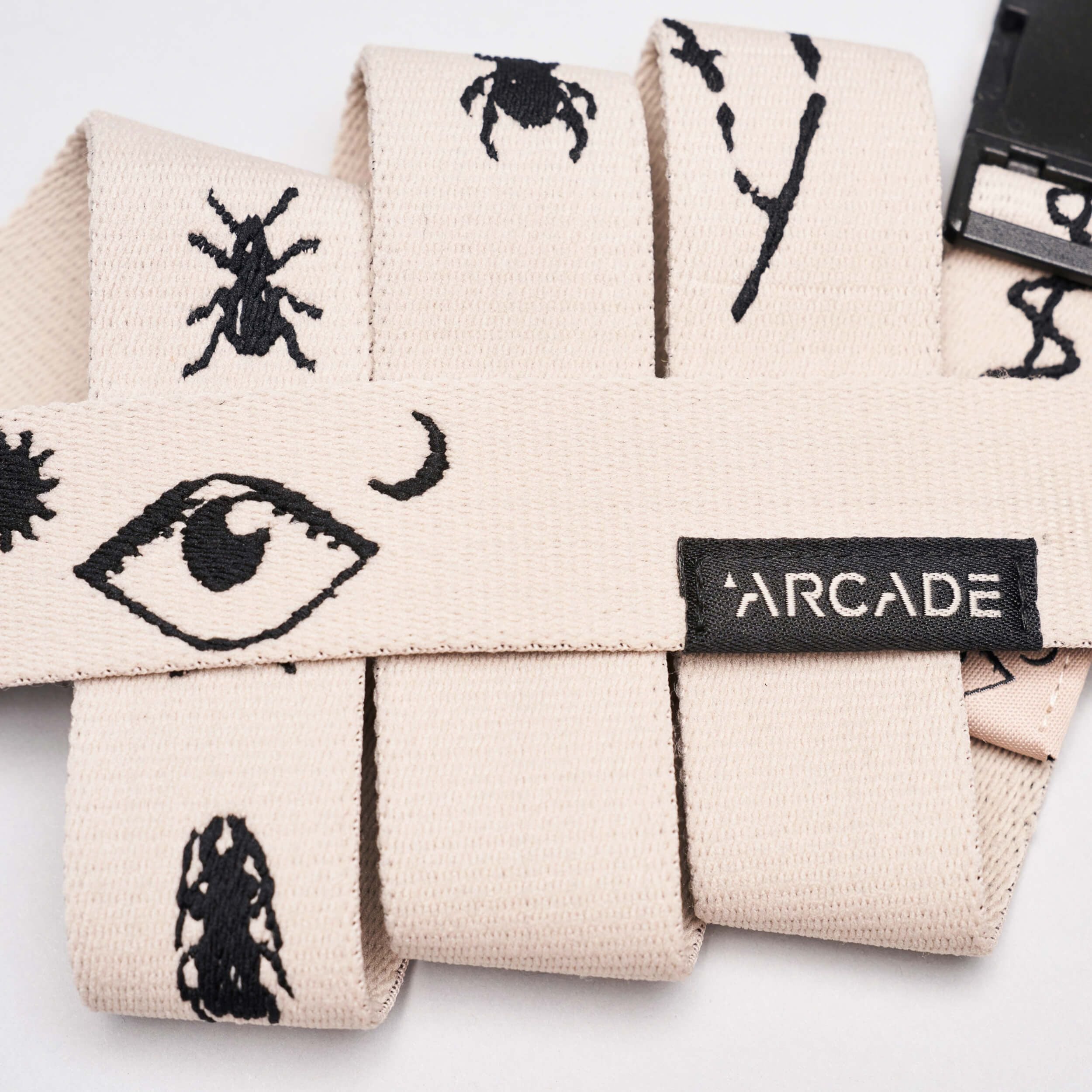 The Charmer Slim by Arcade Belts is a beige stretch webbing belt with black embroidered eye, insect, and abstract designs, finished with a black buckle and ARCADE label—perfect for Arcade Belts fans or anyone seeking a reliable travel belt.