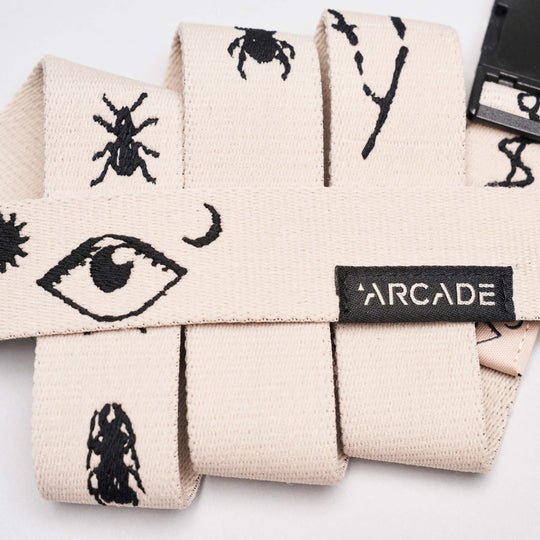 The Charmer Slim by Arcade Belts is a beige stretch webbing belt with black embroidered eye, insect, and abstract designs, finished with a black buckle and ARCADE label—perfect for Arcade Belts fans or anyone seeking a reliable travel belt.