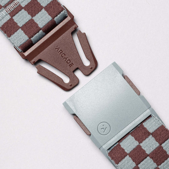 Close-up of the Arcade Belts Check elastic stretch belt in a brown and gray checkered pattern, featuring a low-profile plastic buckle with ARCADE branding and a silver clasp with a smiley face logo, set against a light background.