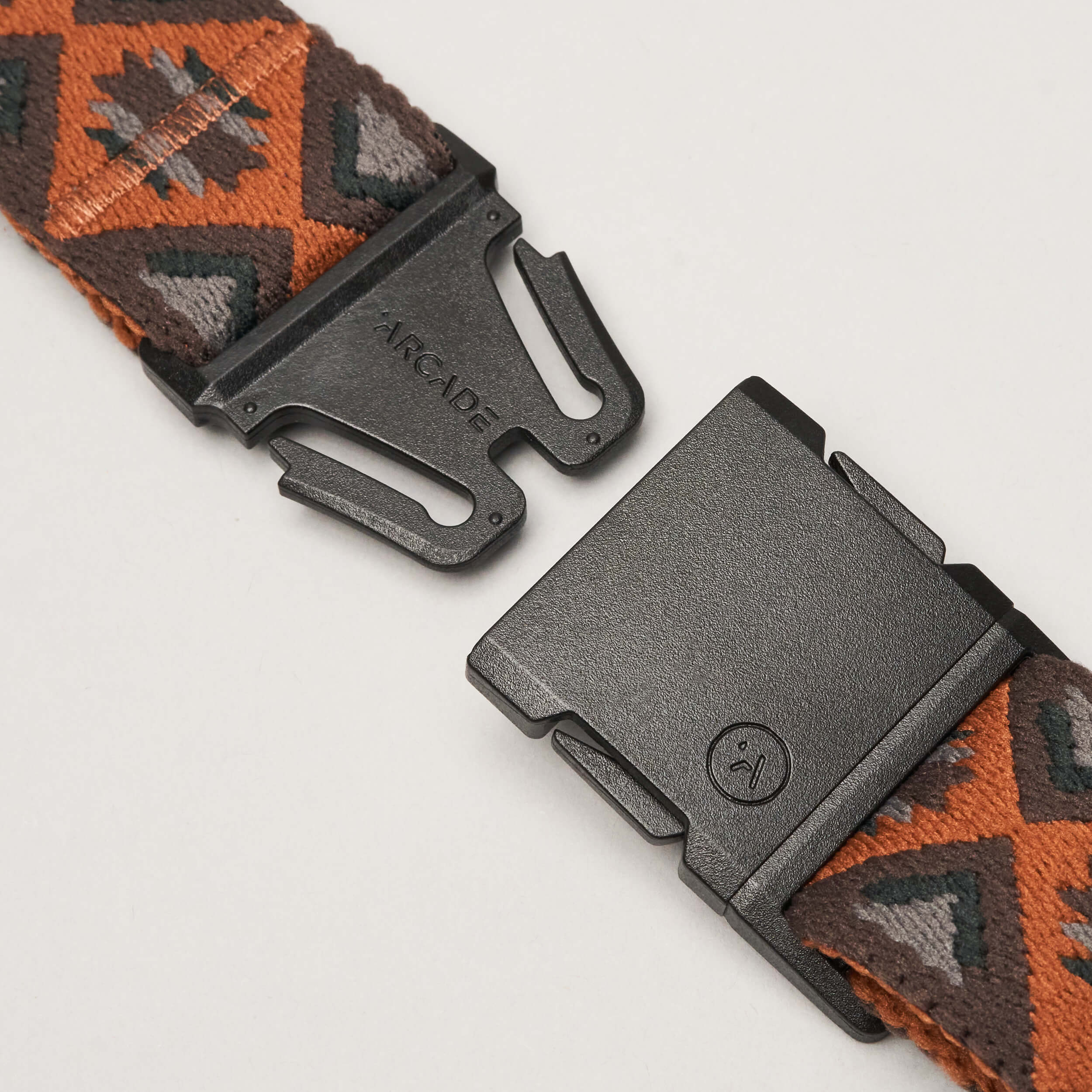 A close-up of the black plastic buckle on the Arcade Belts Vernan Kee elastic stretch belt, featuring brown, orange, and gray geometric designs. The open buckle displays both ends against a plain light background.