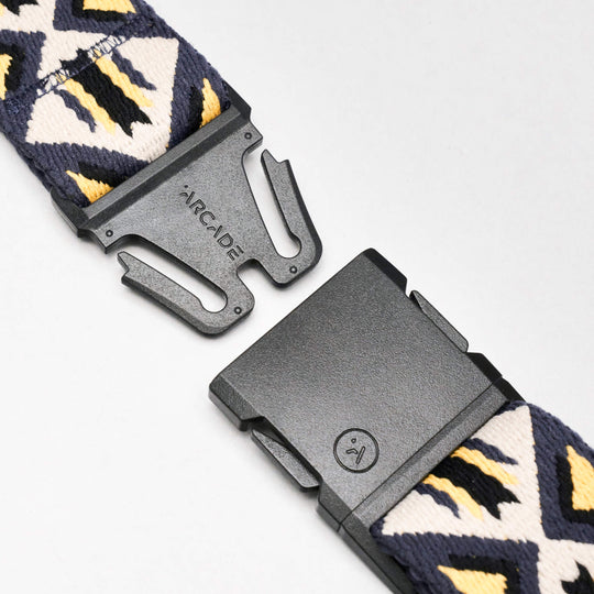 A close-up of the Vernan Kee belt from Arcade Belts shows a black ARCADE-branded buckle and geometric patterns in navy, white, and yellow, inspired by traditional rugs.