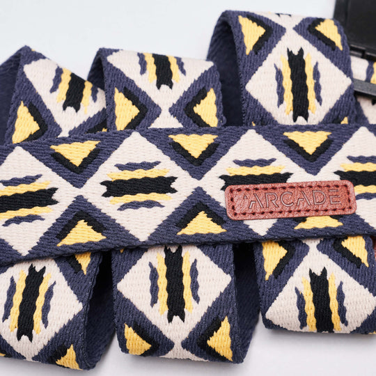 A close-up of the Vernan Kee fabric strap by Arcade Belts shows geometric shapes inspired by traditional rug patterns in navy blue, yellow, and white, with a brown leather tag featuring the brand’s name.