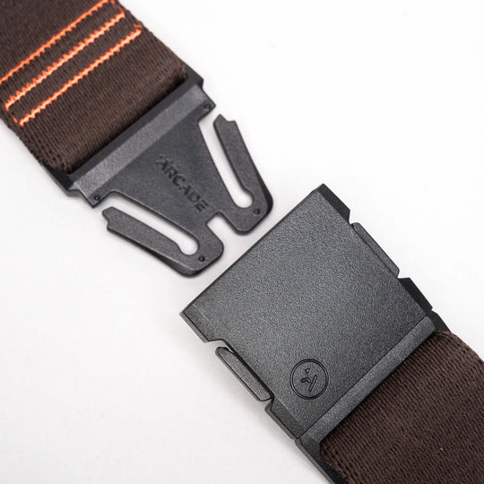 A close-up of the Arcade Belts DDC: a black plastic buckle with logo, attached to a brown stretch webbing strap with orange stripes, shown on a plain white background.