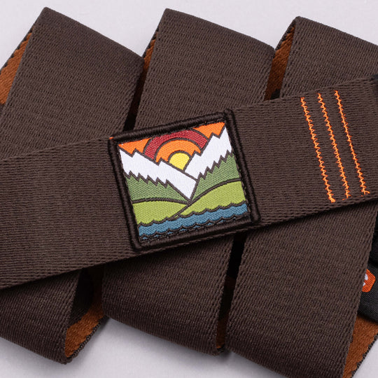 A close-up of the Arcade Belts DDC: a brown stretch webbing strap with orange stitching and a vibrant patch depicting stylized mountains, a river, green fields, and a setting sun.