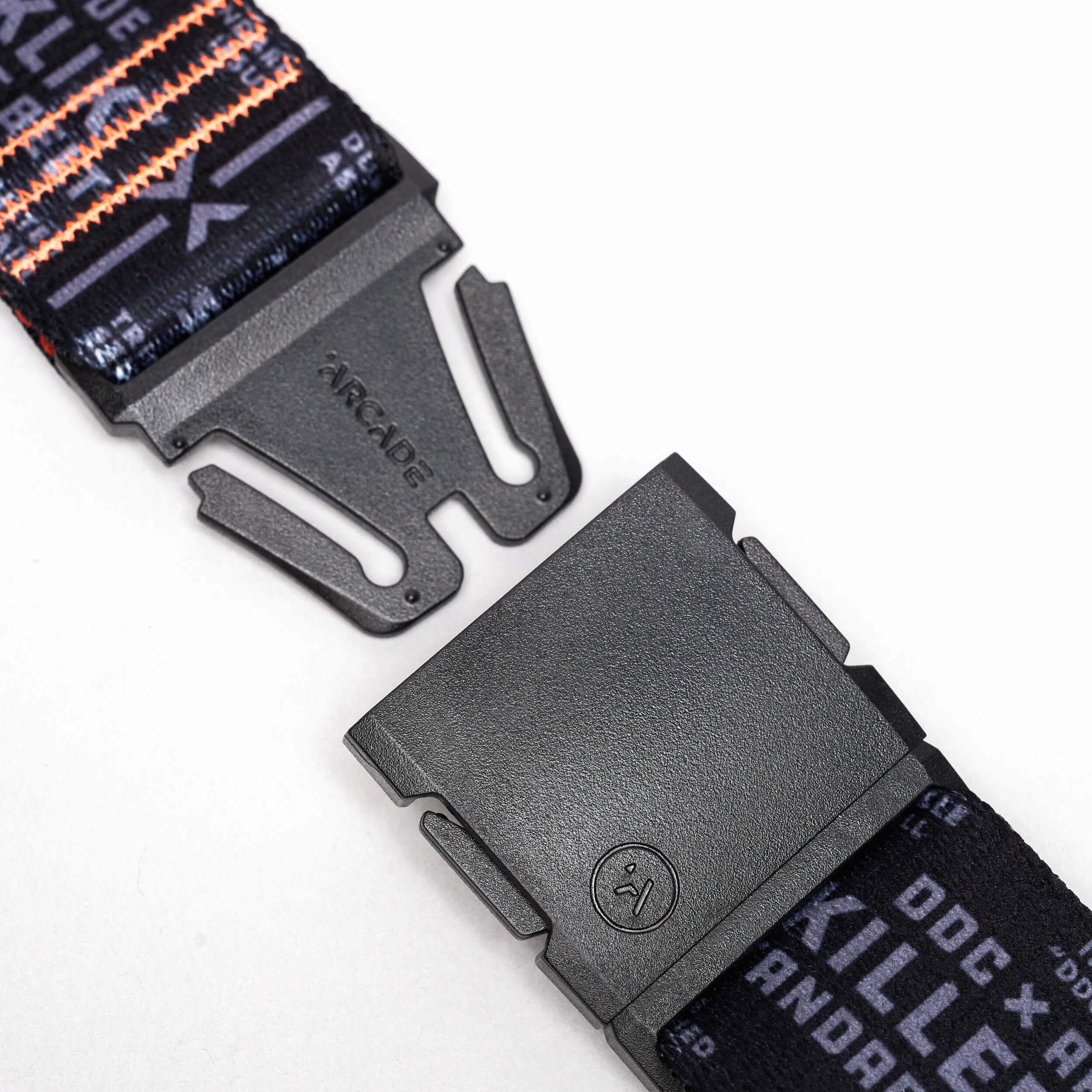 Close-up of a black buckle on an Arcade Belts DDC strap, featuring the word ARCADE and other text, with the elastic stretch belt partially open against a white background.