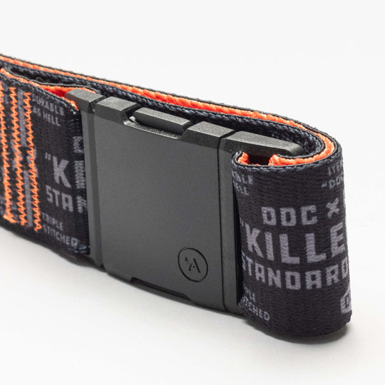 A close-up of the black DDC Low Profile Belt by Arcade Belts, featuring orange stitching, a rectangular black buckle, and white printed text with the words DOC, KILLER, and STANDARD—designed for travel.