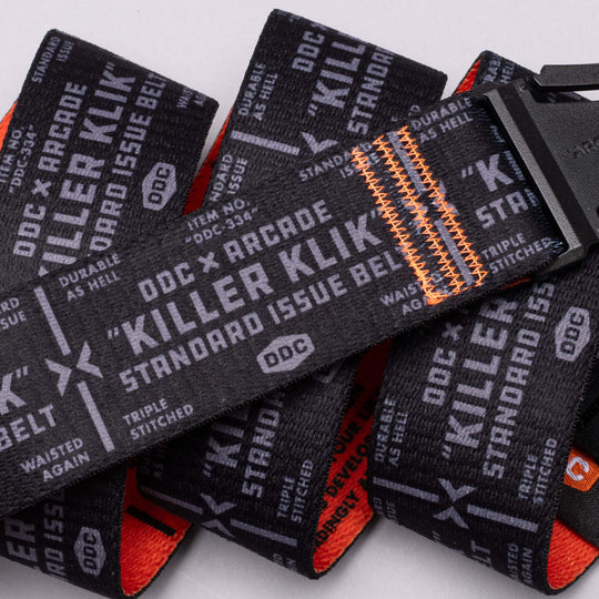 A low-profile Arcade Belts DDC belt with white Killer Klik Standard Issue Belt text, stretch webbing, triple orange stitching, and a black plastic buckle is coiled on a white surface.