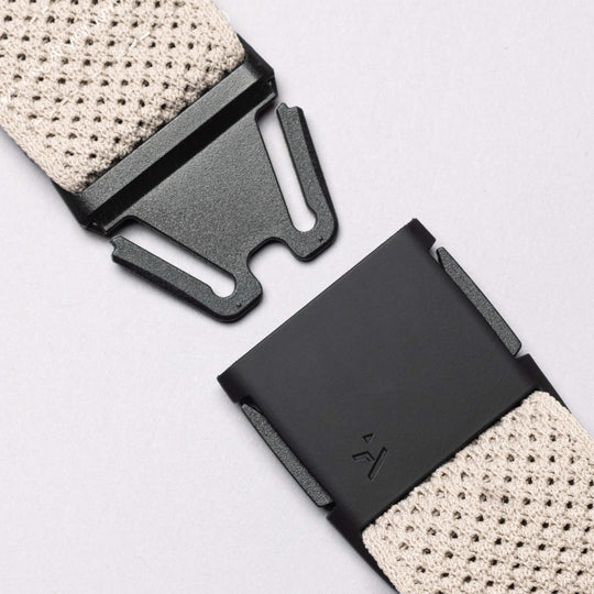 Close-up of the Arcade Belts Momentum: a black metal buckle with a modern geometric design on a beige textured strap—ideal for travel or low-profile wear—set against a neutral background.