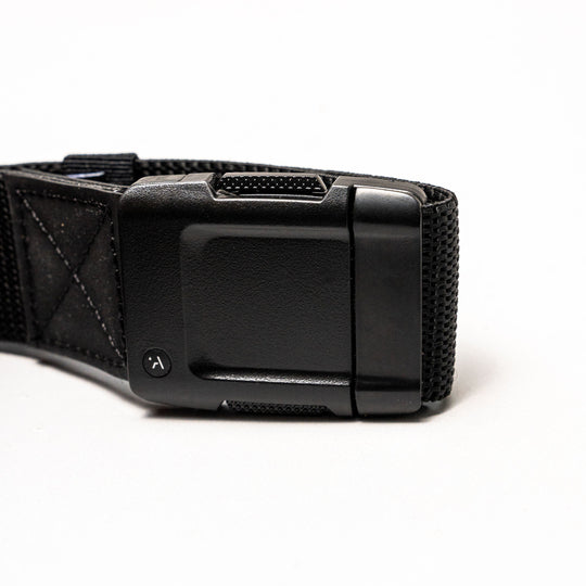 Close-up of the Arcade Belts Motion: a black fabric strap with a rectangular, textured plastic buckle on a white background. The buckle has a small round button with a curved arrow for secure fastening and easy adjustment—perfect for travel.