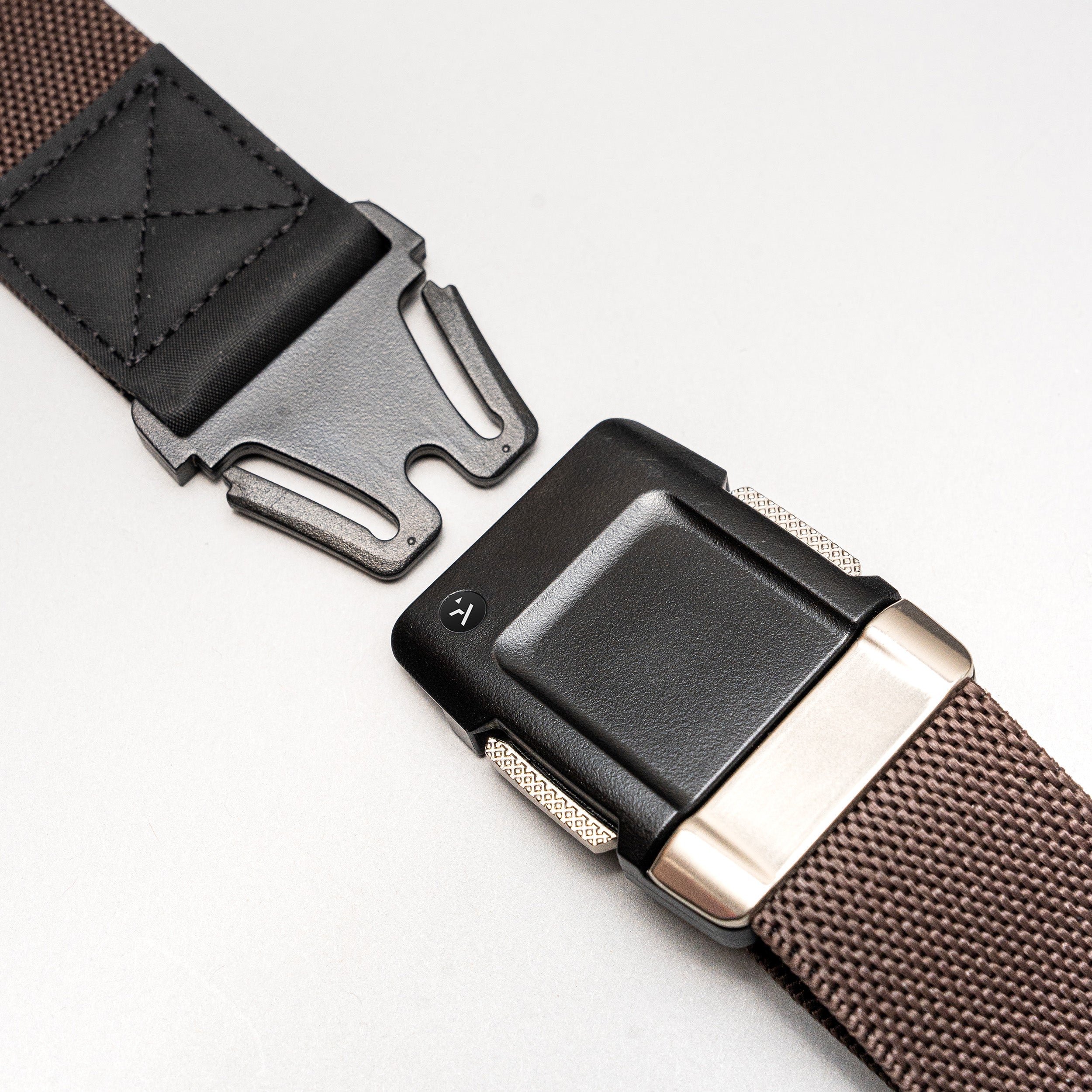 Close-up of the Arcade Belts Motion brown woven low-profile belt with a black and silver buckle and leather detail near the buckle, shown partially unfastened on a light grey background.