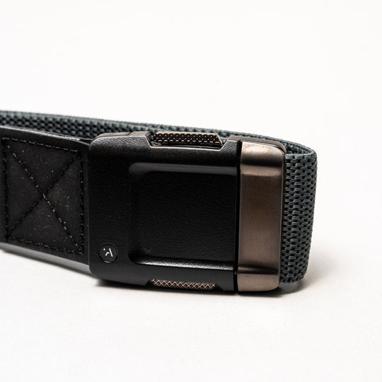 A close-up of the Arcade Belts Motion belt features a sturdy, rectangular matte black buckle and a grey woven elastic stretch strap, set against a plain white background.