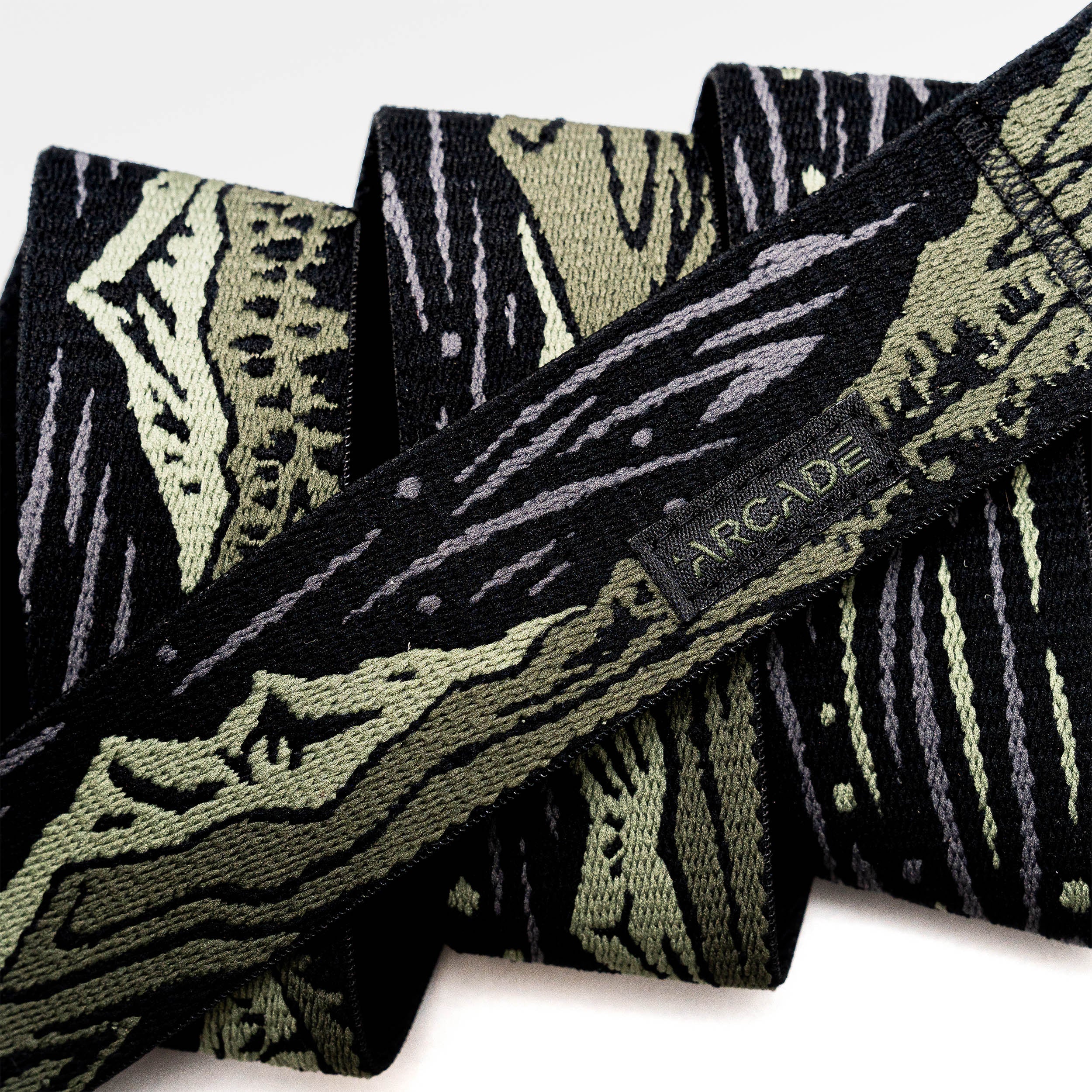 A close-up of the Arcade Belts Out of Range fabric belt, showcasing a black and olive green camo pattern, a small black tag with the brand name stitched on it, and a sturdy plastic buckle.