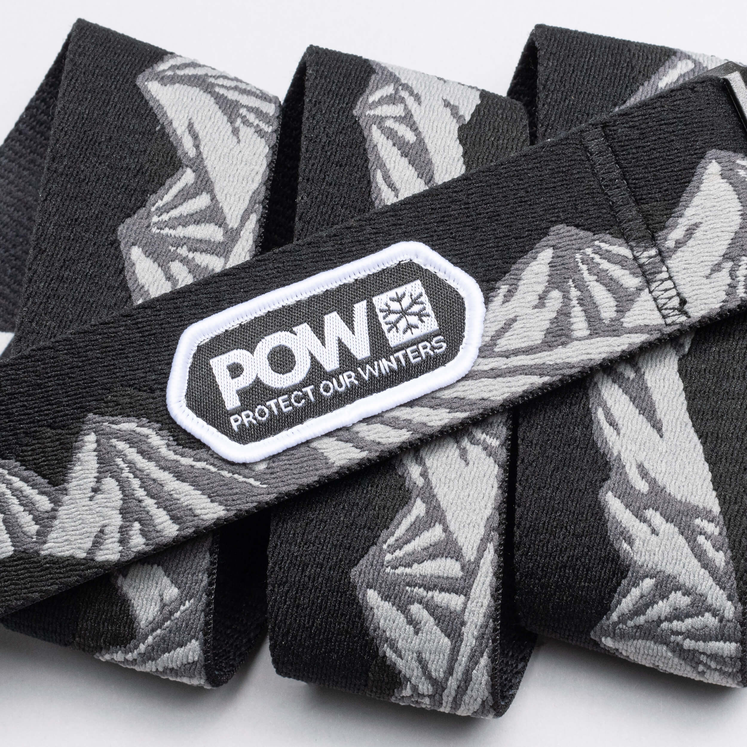 The Arcade Belts POW x Joseph Toney is a black and gray stretch webbing belt with mountain patterns, a low profile design, and a white POW PROTECT OUR WINTERS patch—showcasing classic Arcade Belts style.
