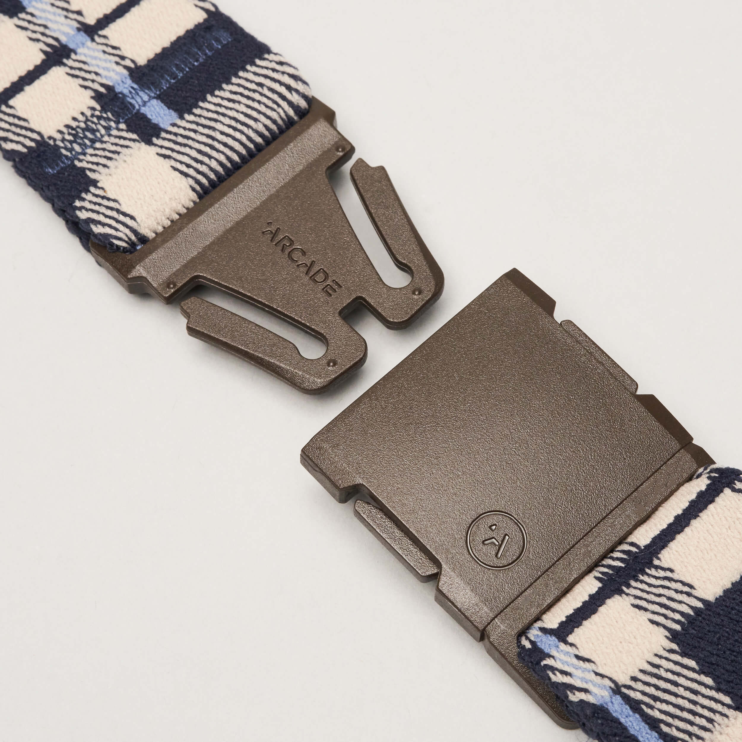 A close-up of the Plaid Elastic Stretch Belt by Arcade Belts features a blue and cream plaid strap and an unfastened brown plastic buckle showing the Arcade logo and name.