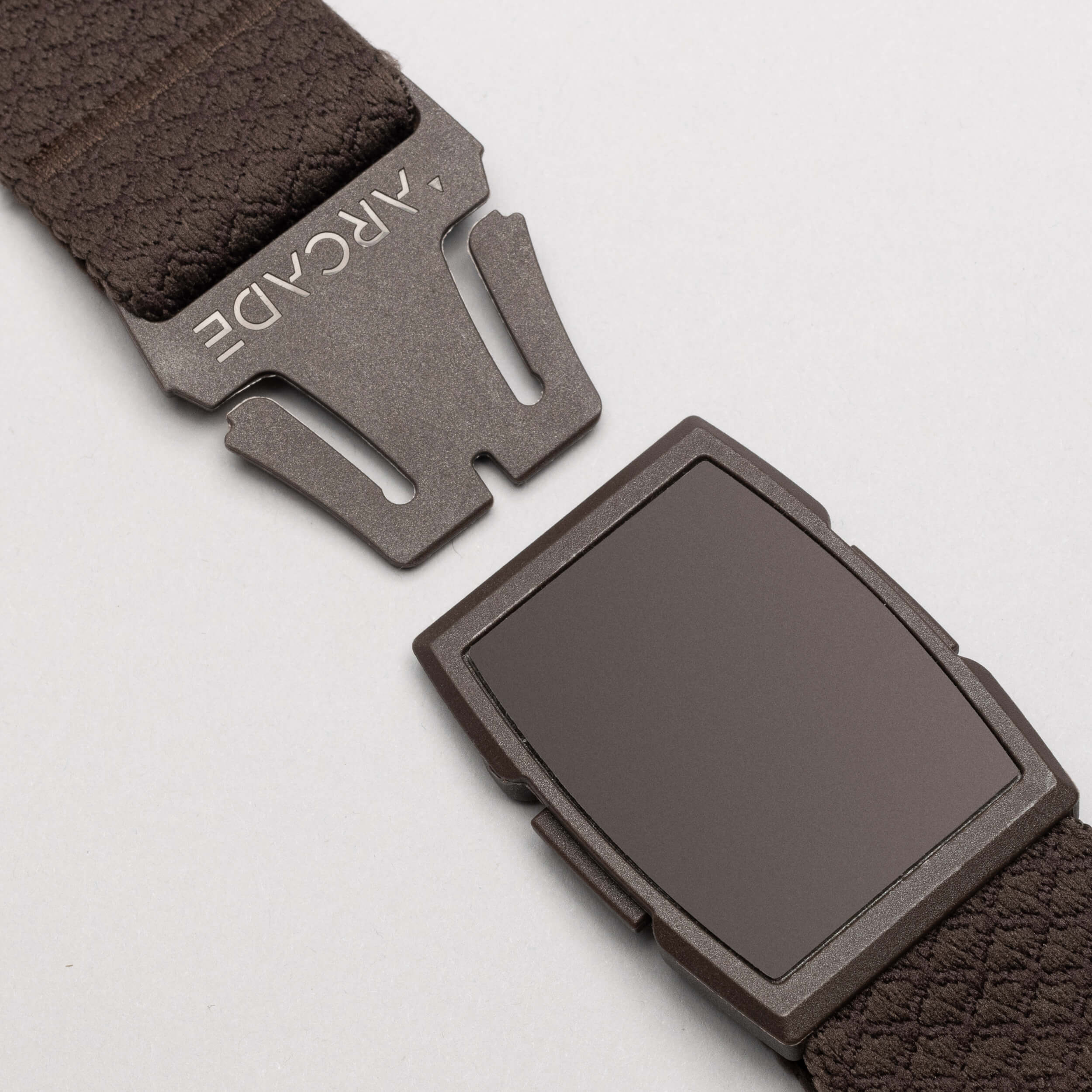 Close-up of the Salient belt by Arcade Belts, featuring a brown low-profile design with a matte rectangular magnetic buckle engraved with ARCADE. The partially unclasped belt is set against a plain light grey background.