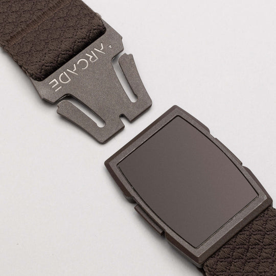 Close-up of the Salient belt by Arcade Belts, featuring a brown low-profile design with a matte rectangular magnetic buckle engraved with ARCADE. The partially unclasped belt is set against a plain light grey background.