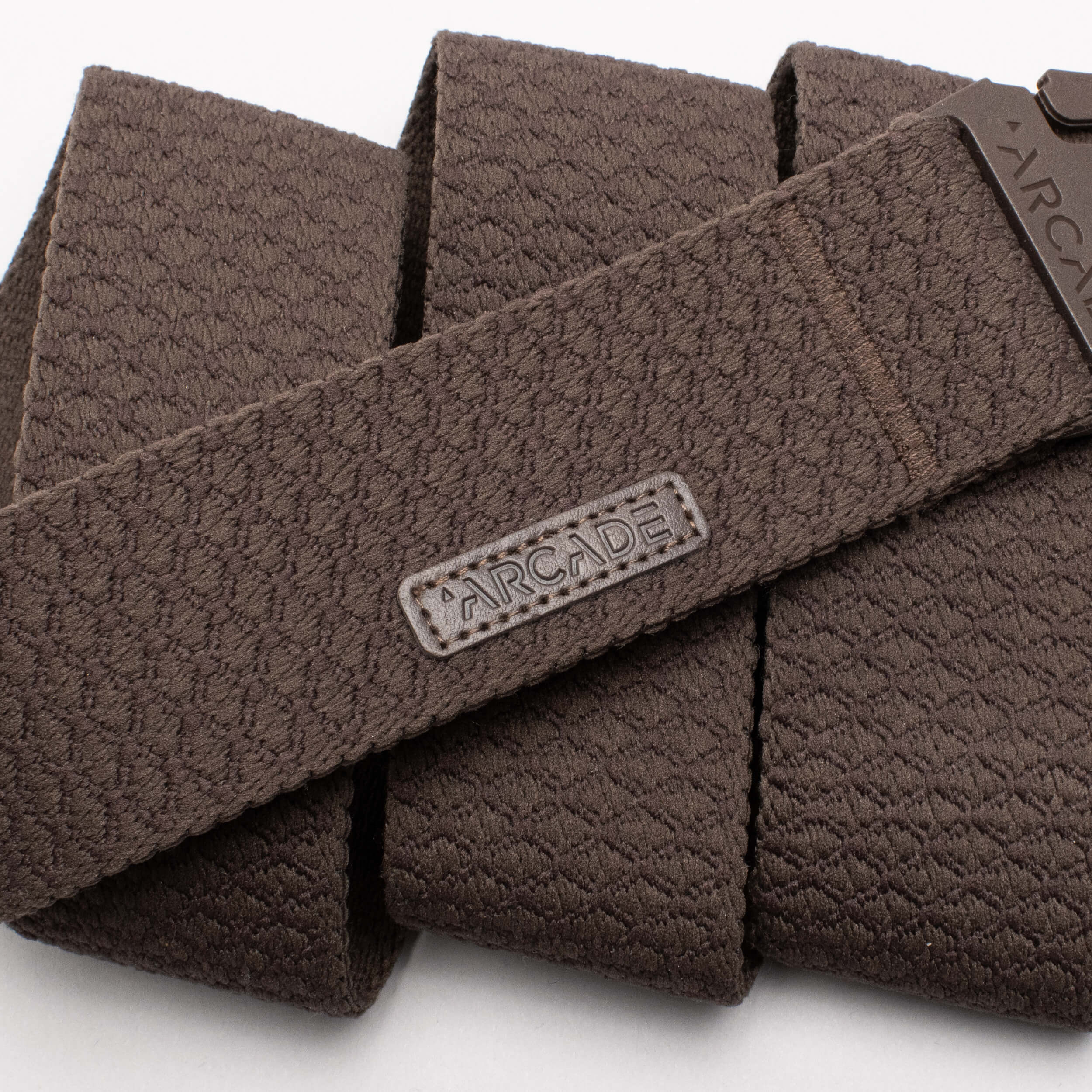 Close-up of the Salient belt by Arcade Belts, featuring a textured brown geometric pattern, low profile buckle, flexible stretch design, and stitched ARCADE label—perfect for travel or daily wear.