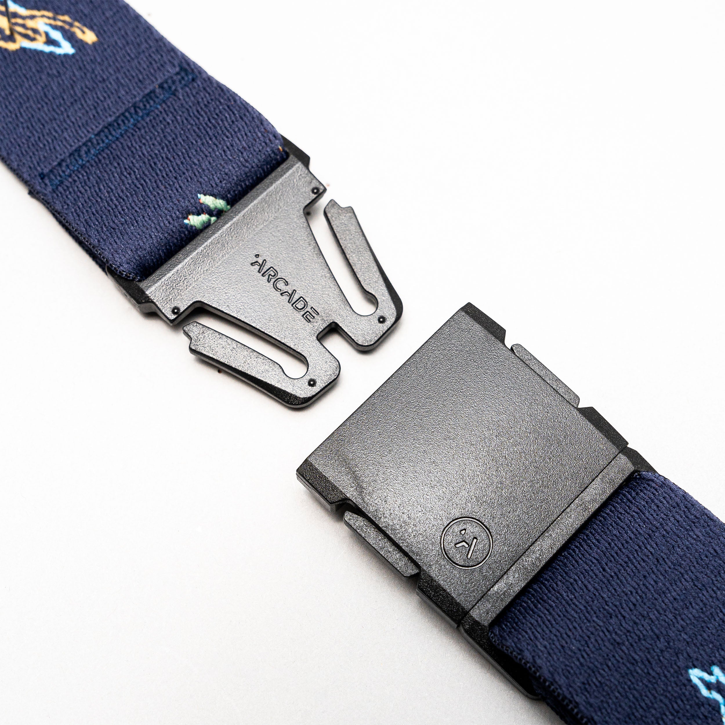 A close-up of the Arcade Belts Save the Waves navy blue stretch belt with a black plastic buckle, partially unfastened. The ARCADE logo is visible on the buckle; perfect for travel belt enthusiasts.