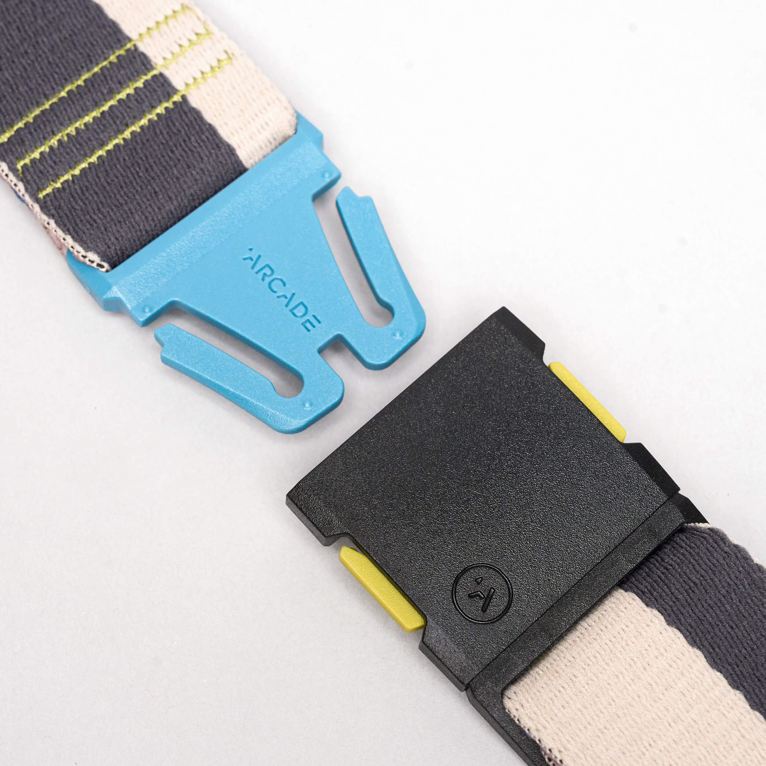 A close-up of a blue plastic buckle labeled ARCADE, attached to the Arcade Belts Sierra elastic stretch belt with striped grey, cream, and yellow-green fabric.