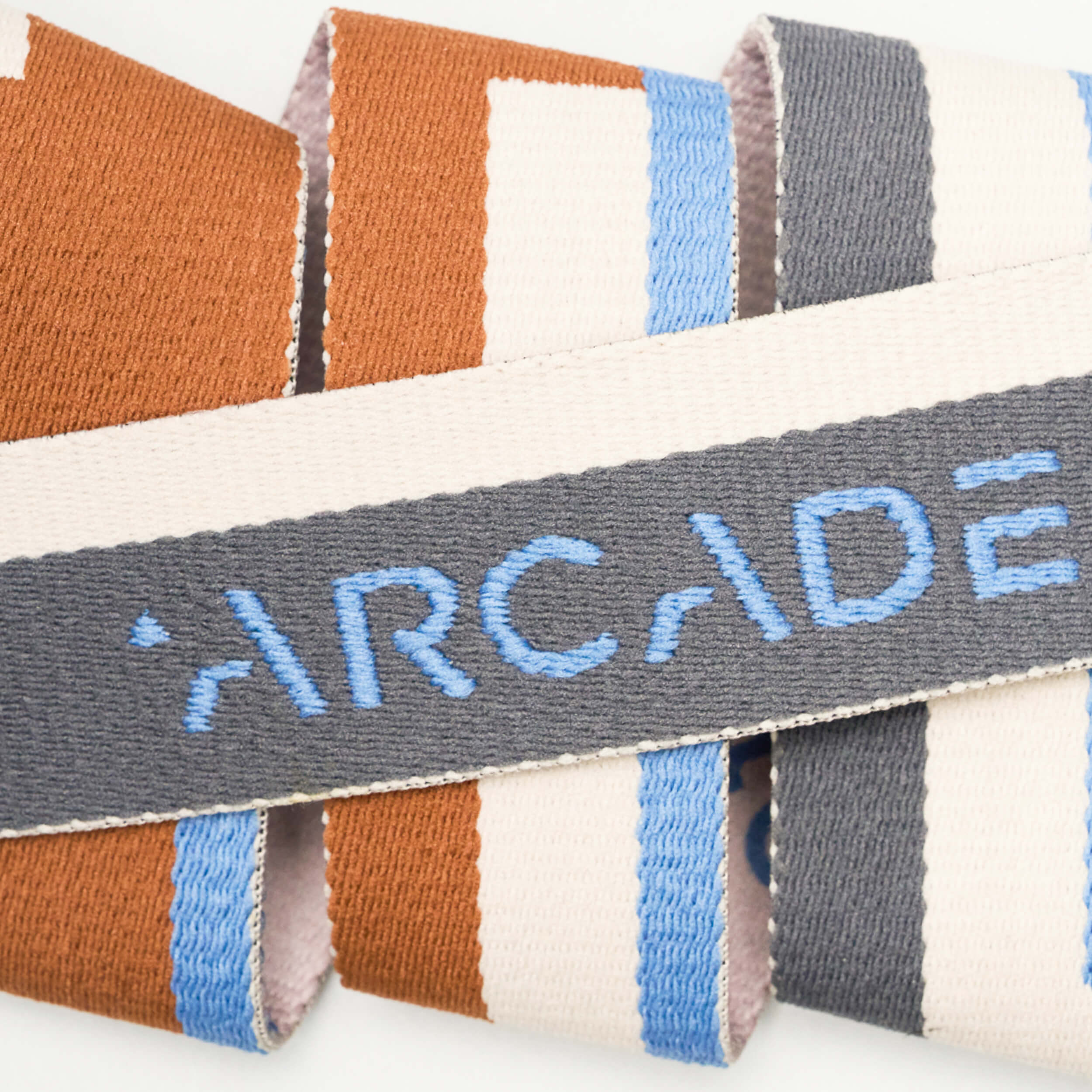 A close-up of the Sierra low profile belt by Arcade Belts, featuring ARCADE embroidered in blue on gray, with brown, white, blue, and gray stripes for a comfortable and stylish look.