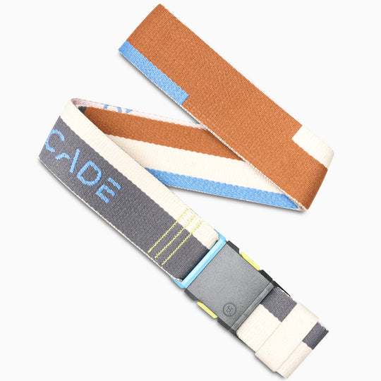 The Arcade Belts Sierra is a fabric travel belt with brown, white, gray, and blue color blocks, stretch webbing for comfort, a rectangular gray plastic buckle, and ARCADE partly visible in light blue.