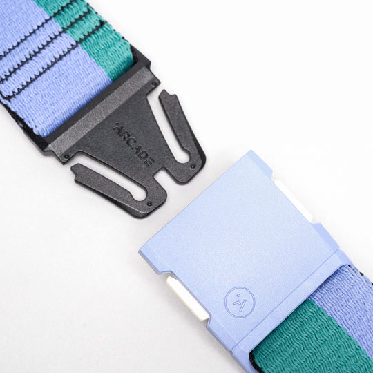 A close-up of the Arcade Belts Sierra travel belt shows a black plastic buckle with a small smiley face icon and a light blue magnetic clasp. The stretch webbing features woven stripes in purple, green, and black.