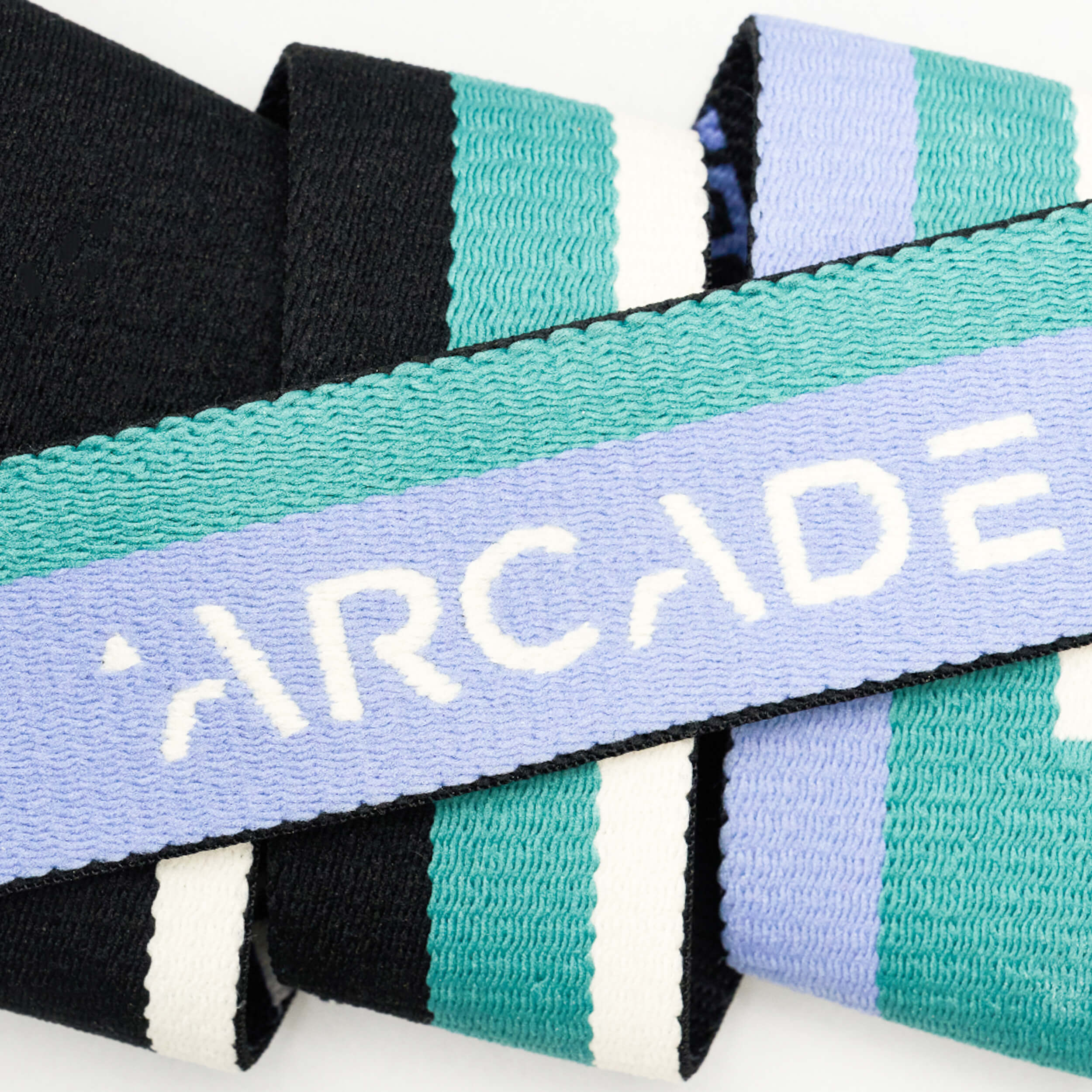 A close-up of the Sierra strap by Arcade Belts, featuring teal, white, and lavender stripes with ARCADE in white on the lavender stripe, highlights the brand’s signature stretch webbing.