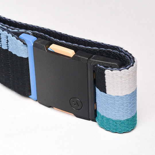 A close-up of the Arcade Belts Sierra, a low-profile belt with blue, white, black, and teal stretch webbing stripes. It has a black rectangular buckle and a small circular logo, shown coiled on a plain white background.