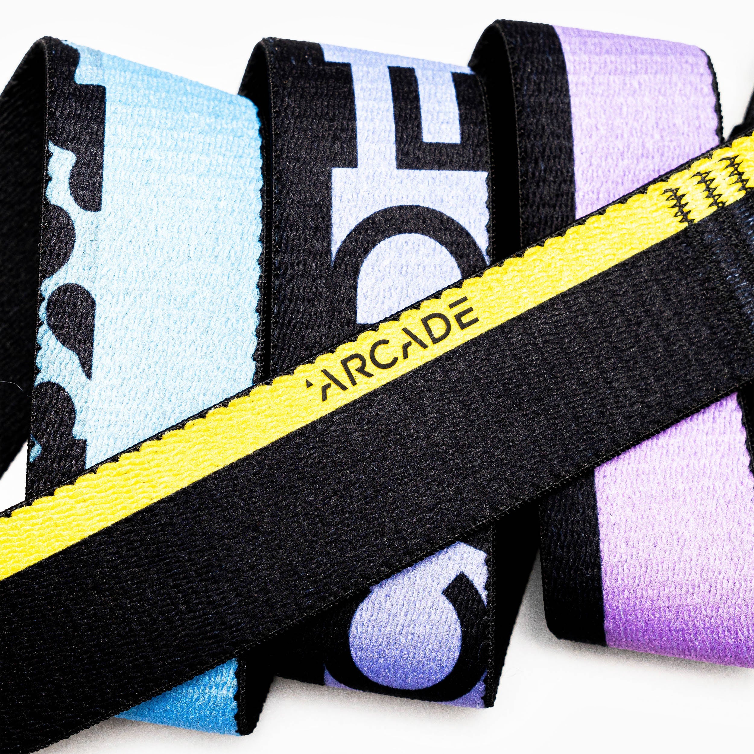 A close-up of Sierra elastic stretch belts by Arcade Belts, featuring a black band with yellow stripe and logo, intertwined with blue, purple, and black patterned bands, all equipped with sturdy plastic buckles.
