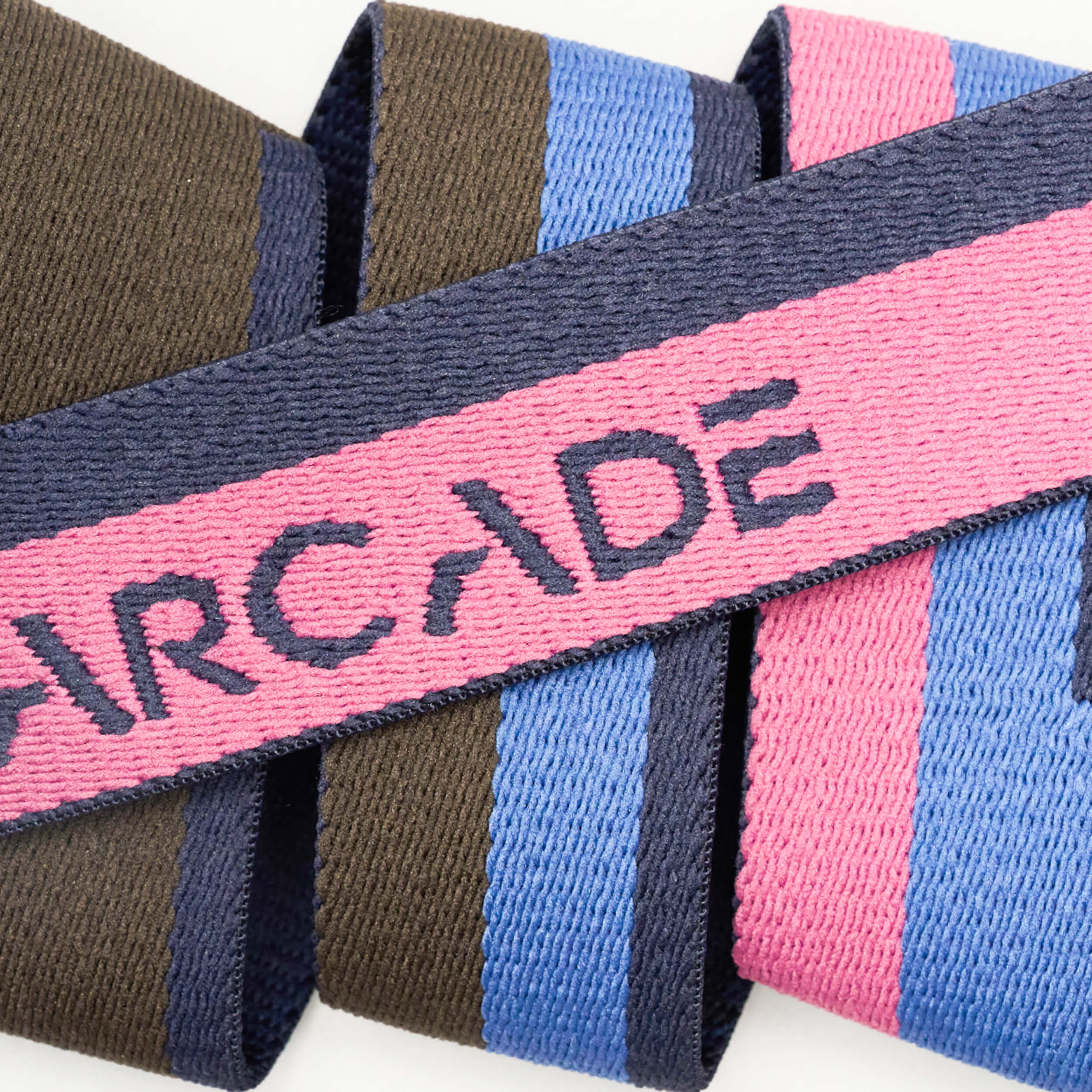 A close-up of the Arcade Belts Sierra, an elastic stretch belt with navy, olive, pink, and blue stripes. ARCADE is stitched in navy on the pink section, highlighting this stylish travel belt design.