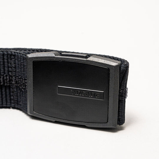 A close-up of the Arcade Belts Stamped, a low-profile belt with a rectangular black plastic buckle embossed with ARCADE. Its stretch webbing ensures comfort. The product is shown against a plain white background.