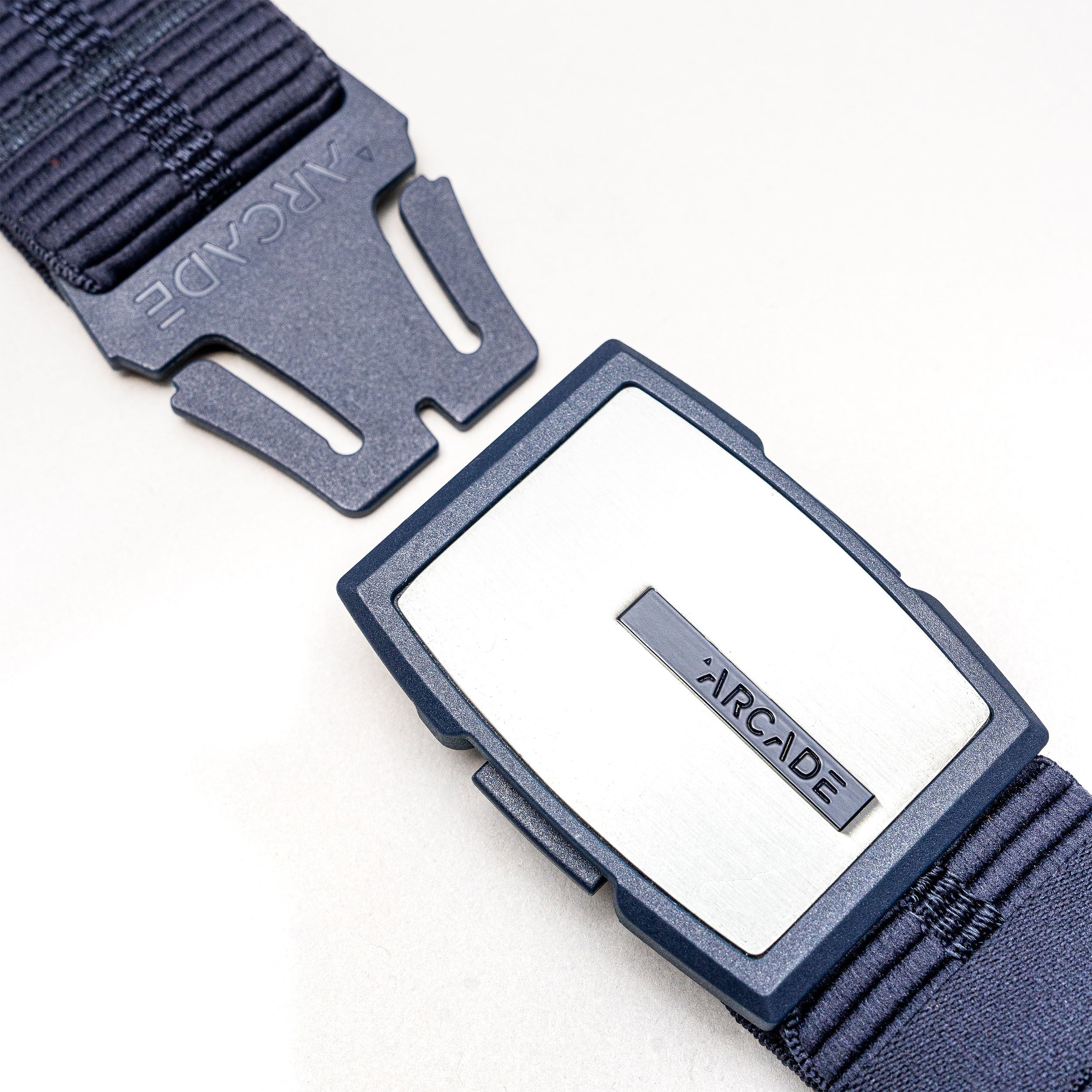 A close-up of the Arcade Belts Stamped navy blue stretch webbing belt with a rectangular silver buckle and ARCADE logo, unbuckled and displayed on a light surface.