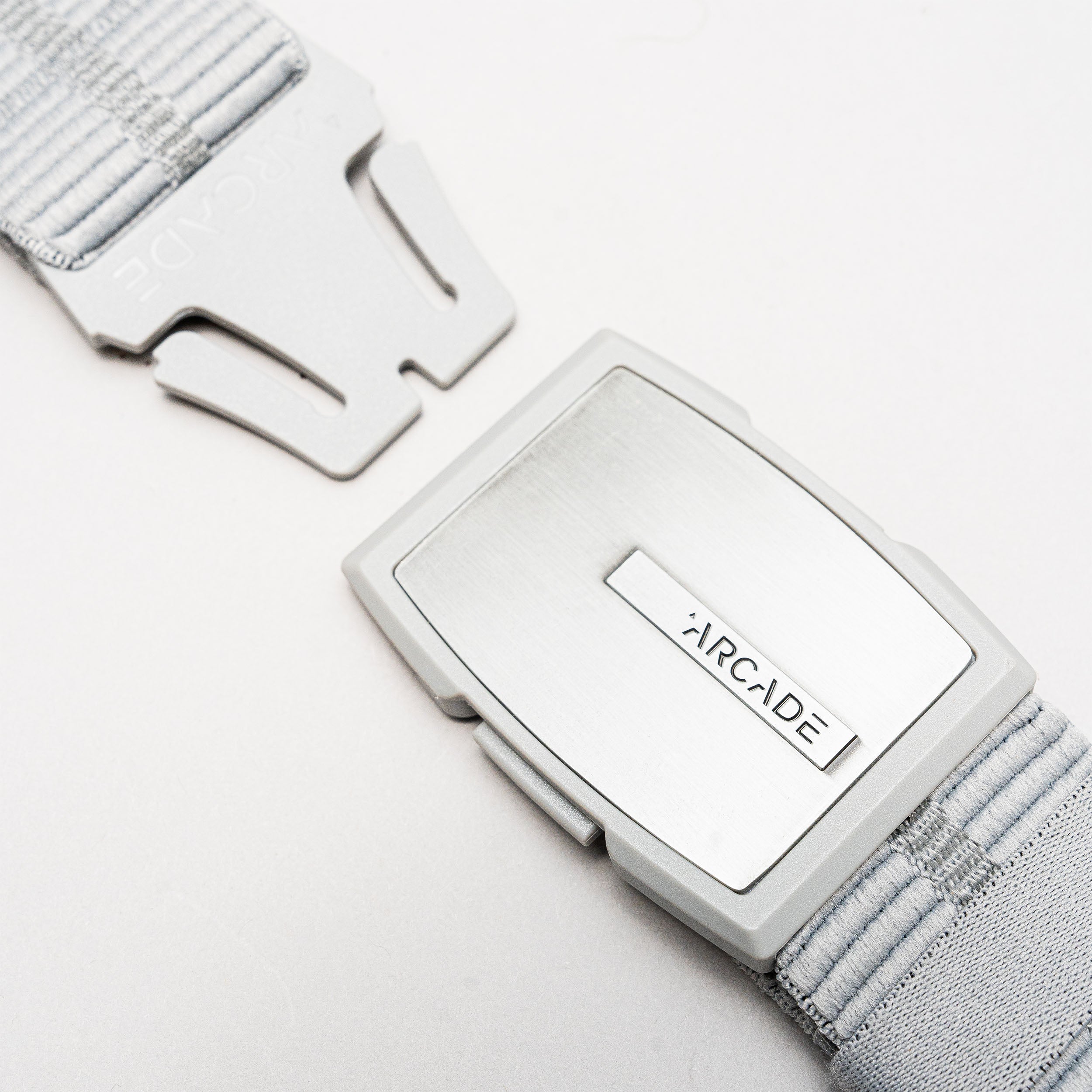 A close-up of the Stamped belt by Arcade Belts shows its light gray, textured elastic strap and sleek rectangular metal buckle. The low-profile buckle features clean lines, making this travel belt comfortable, stylish, and partially unclasped.