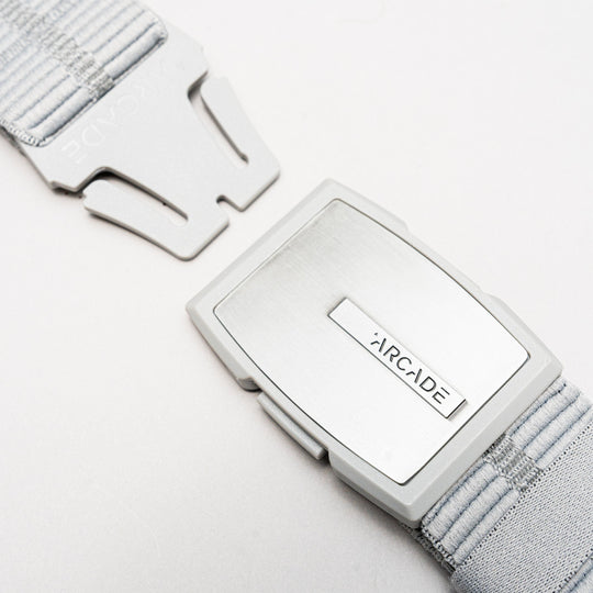 A close-up of the Stamped belt by Arcade Belts shows its light gray, textured elastic strap and sleek rectangular metal buckle. The low-profile buckle features clean lines, making this travel belt comfortable, stylish, and partially unclasped.