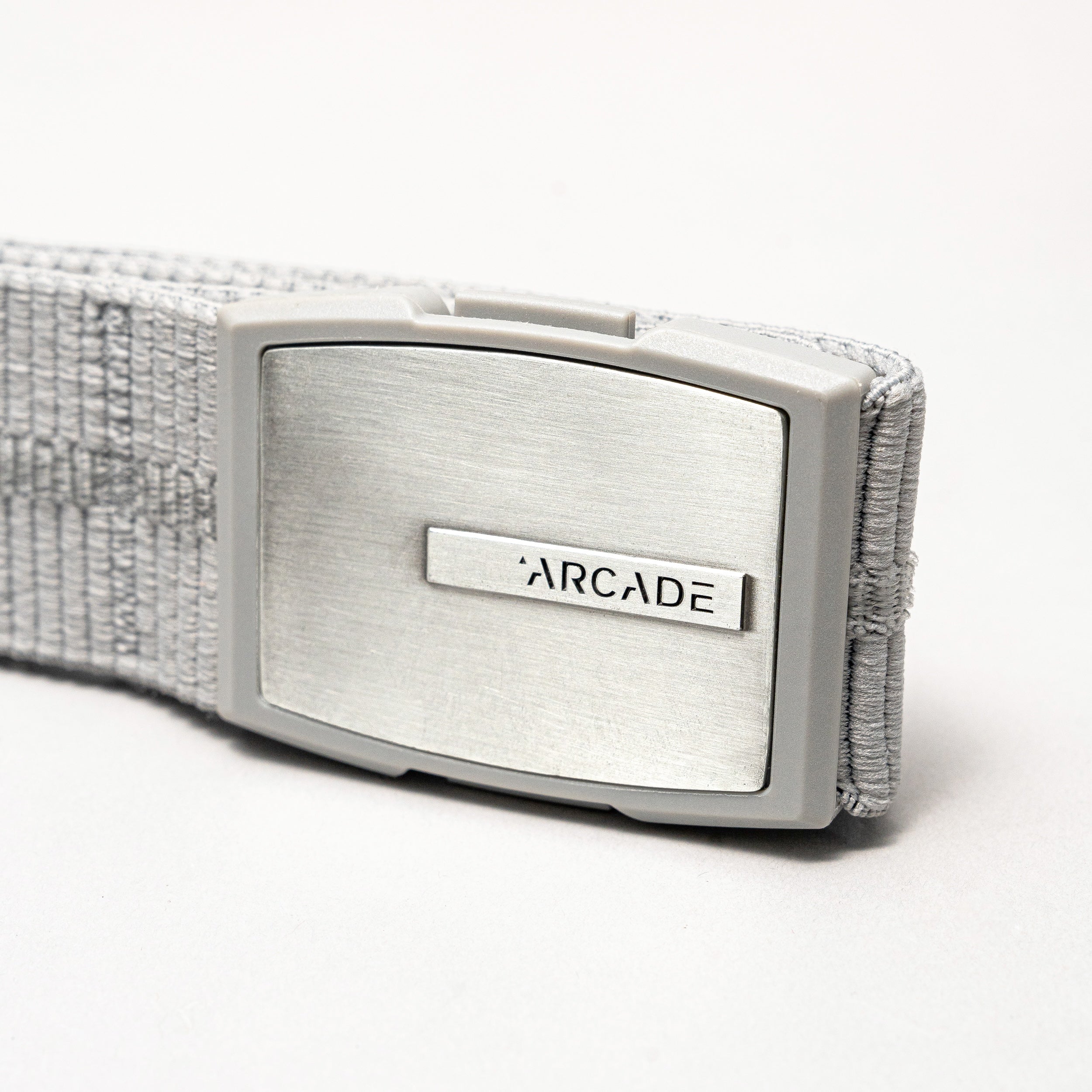 A close-up of the silver Stamped buckle, engraved with ARCADE and attached to a textured light gray elastic stretch belt by Arcade Belts.
