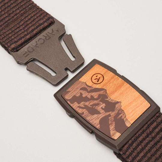 A close-up of the Arcade Belts Woody shows its brown woven design and partially open metal clasp with a wooden inlay engraved with a mountain scene and a smiley face. The low-profile Woody belt seamlessly blends style and function.